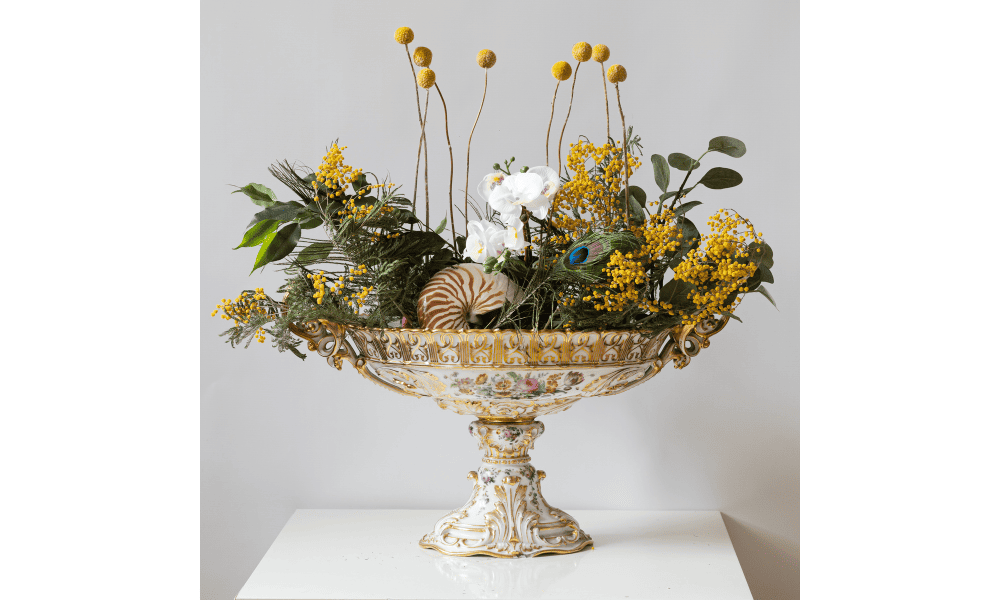 A decorative porcelain bowl filled with a vibrant floral arrangement, featuring yellow flowers, greenery, and a peacock feather, placed on a white table against a plain background.