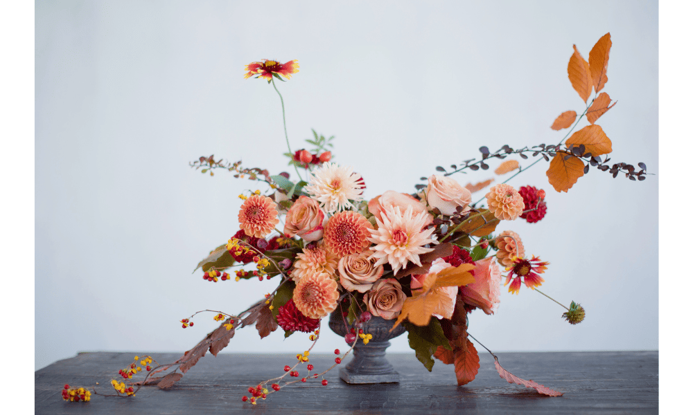 Elegant floral arrangement in a vintage vase with a mix of autumnal flowers in shades of orange, pink, and red, set on a dark wooden table against a light blue background