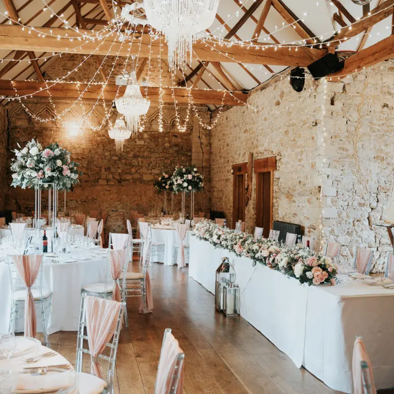 Indoor wedding reception with round tables covered in pink tablecloths and decorated with glassware, fairy lights and floral centrepieces.