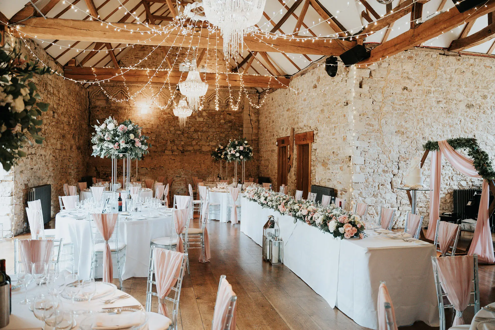 Indoor wedding reception with round tables covered in pink tablecloths and decorated with glassware, fairy lights and floral centrepieces.