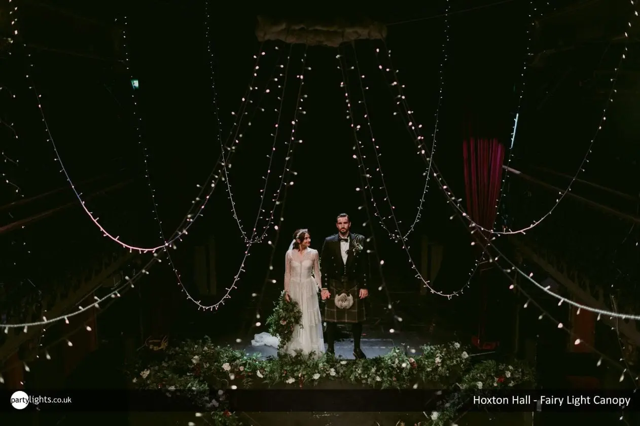 Married couple stood on a stage decorated with draping fairy lights