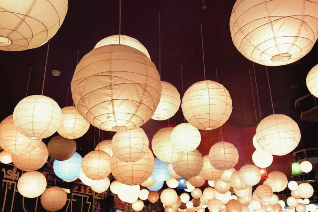 Cluster of glowing paper lanterns of various sizes hanging from the ceiling, creating a warm and ambient lighting effect.