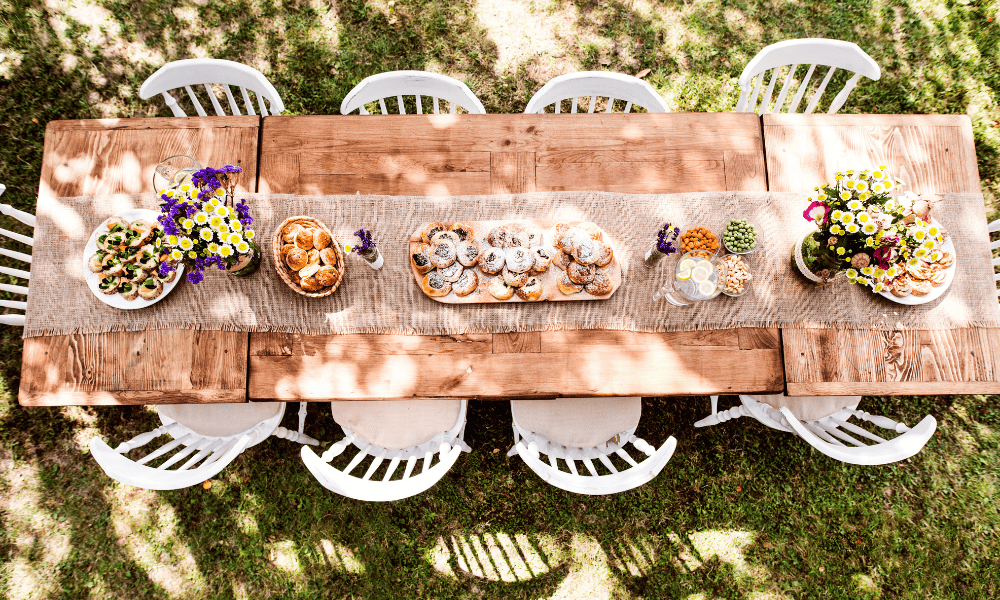 Overhead view of an outdoor wooden dining table set with pastries, flowers, and snacks. The table is surrounded by white chairs, and it is situated in a shaded garden setting.