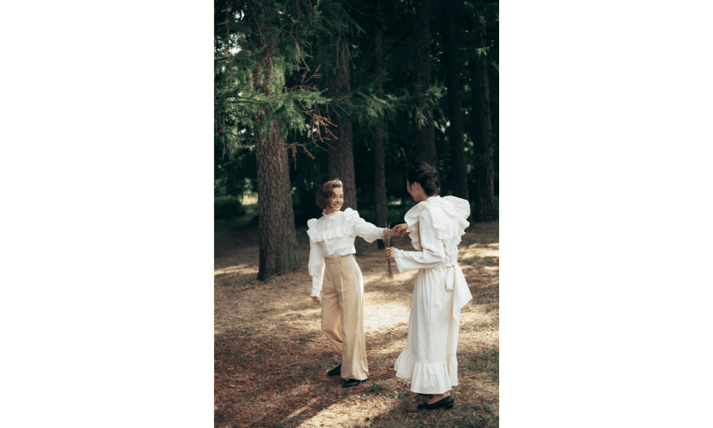 Two women dressed in white, flowy vintage-style outfits, walking and interacting with each other amidst a pine forest setting, with dappled sunlight filtering through the trees