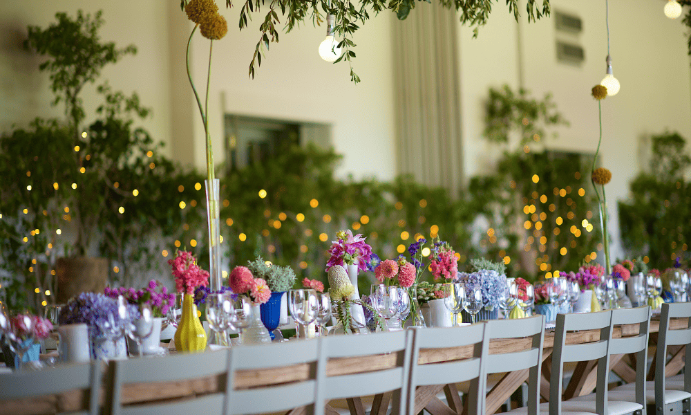 A long banquet-style table decorated with colourful floral arrangements and glasses, set up for an indoor event with soft, ambient lighting and greenery in the background.