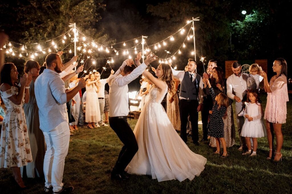 Bride and groom sharing their first dance outdoors at night, surrounded by guests holding sparklers, with string lights illuminating the scene