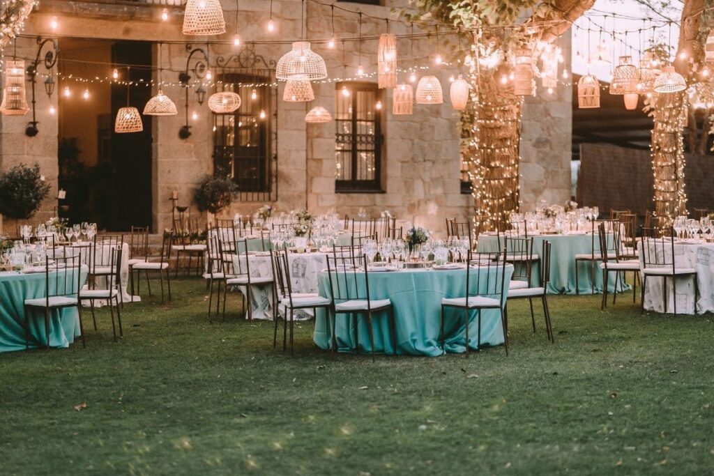 Outdoor wedding reception with round tables, turquoise tablecloths, and hanging lanterns, illuminated by string lights wrapped around trees.