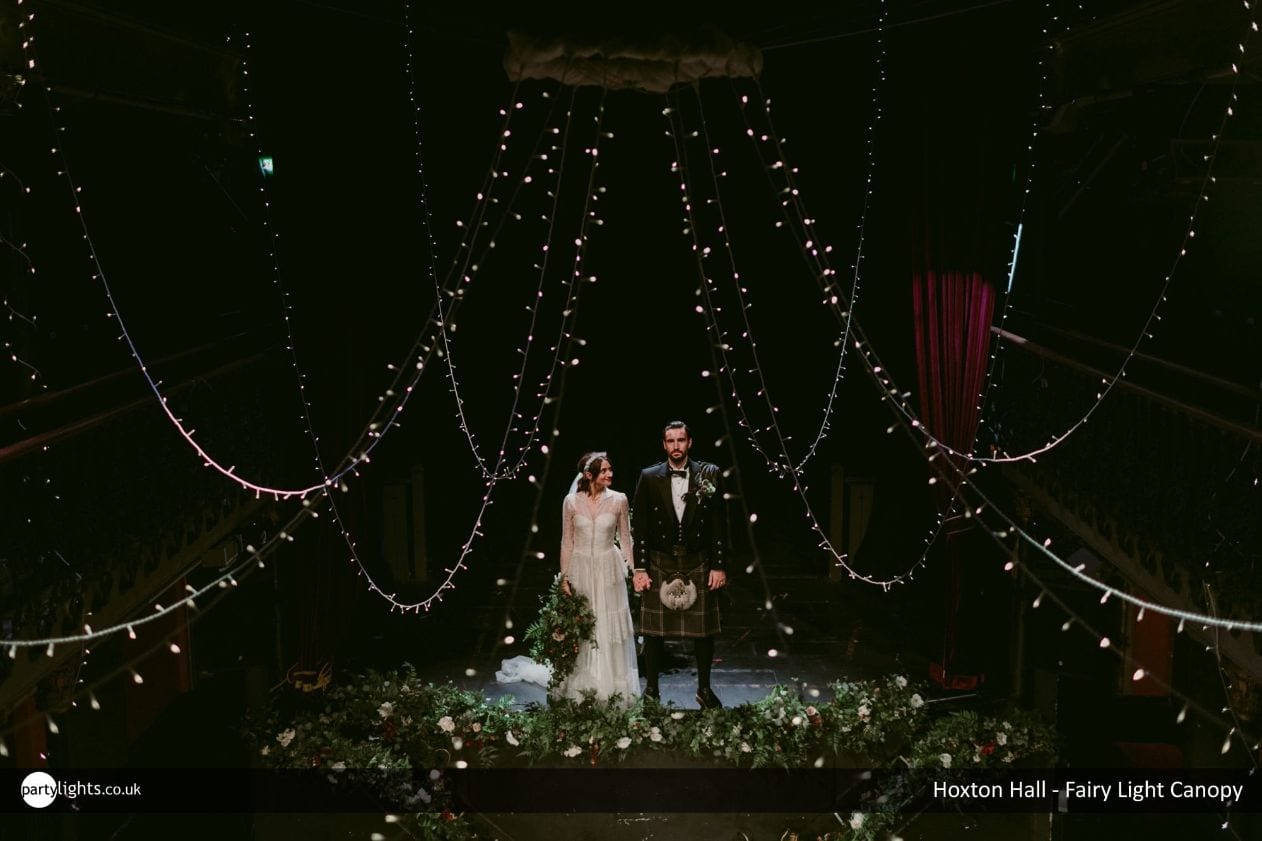 Married couple stood on a stage decorated with draping fairy lights